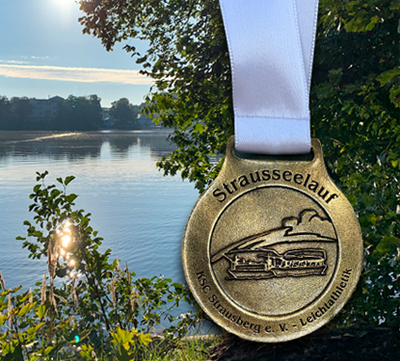 Die verdiente Medaille strahlt im Morgenlicht vor dem idyllischen Straussee – Erinnerung an einen sportlichen und sonnigen Tag beim Strausseelauf 2025.